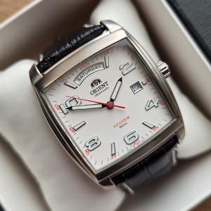 Può includere: Un orologio da polso automatico Orient con cassa rettangolare in titanio, quadrante bianco e cinturino in pelle nera. Il quadrante presenta grandi numeri argentati, una finestrella giorno-data, una lancetta dei secondi rossa e la scritta Orient Automatic, Titanium, 50M.