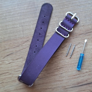 Könnte beinhalten: Ein lila Leder-Uhrenarmband mit silberfarbenen Beschlägen, einschließlich Schnallen und Schlaufen. Zwei silberne Stifte und ein blauer Schraubendreher sind ebenfalls vorhanden. Das Armband liegt auf einer Holzoberfläche.