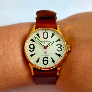Puede incluir: Un reloj de pulsera dorado con una esfera blanca y números negros. El reloj tiene una correa de cuero marrón y dice "PAKETA" y "СДЕЛАНО В СССР" en la esfera.