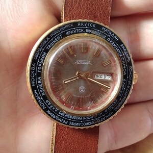 Puede incluir: Un reloj de pulsera dorado con una correa de cuero negro. La esfera del reloj tiene un fondo plateado con marcadores de hora en n&uacute;meros romanos negros y una ventana de fecha que muestra "ПНД 17". La esfera del reloj est&aacute; rodeada por un anillo negro con nombres de ciudades impresos en blanco.