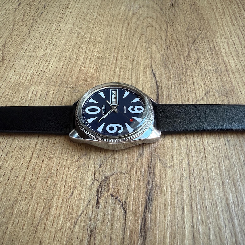 May include: A silver-toned wristwatch with a dark blue face and white numerals. The watch has a black leather band. The watch face displays the day and date. The watch is set against a wooden surface.