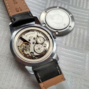 Pu&ograve; includere: Un orologio da polso argentato con un cinturino in pelle nera. Il quadrante dell'orologio &egrave; stato rimosso, rivelando il meccanismo interno dell'orologio. L'orologio &egrave; su una superficie bianca.