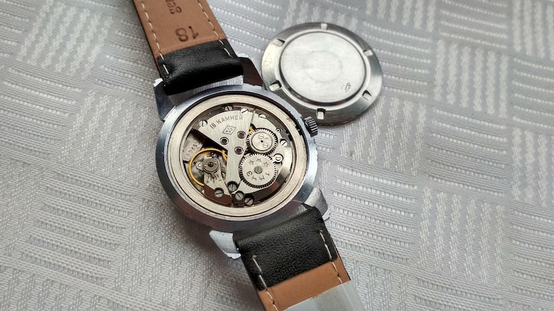 Pu&ograve; includere: Un orologio da polso argentato con un cinturino in pelle nera. Il quadrante dell'orologio &egrave; stato rimosso, rivelando il meccanismo interno dell'orologio. L'orologio &egrave; su una superficie bianca.