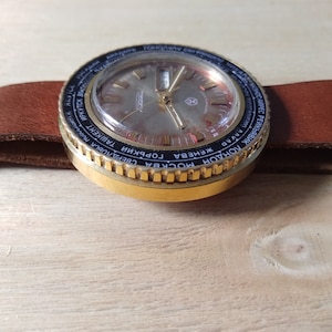 Puede incluir: Un reloj de pulsera dorado con una esfera negra y un bisel giratorio. El bisel est&aacute; marcado con los nombres de ciudades de todo el mundo en escritura cir&iacute;lica. La esfera del reloj tiene una ventana de fecha y un segundero.