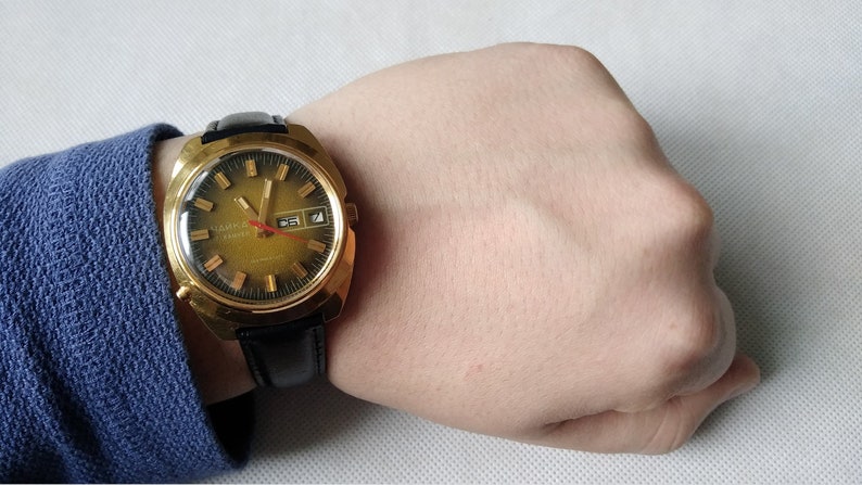 May include: A gold-tone wristwatch with a black leather band. The watch face is round and has a gold-tone bezel. The watch has a date window at the 3 o'clock position and a second hand.