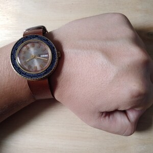Puede incluir: Un reloj de pulsera de cuero marr&oacute;n con una esfera dorada y marcas negras. La esfera del reloj tiene una ventana de fecha y una pantalla de hora mundial. El reloj est&aacute; en la mu&ntilde;eca de una persona.
