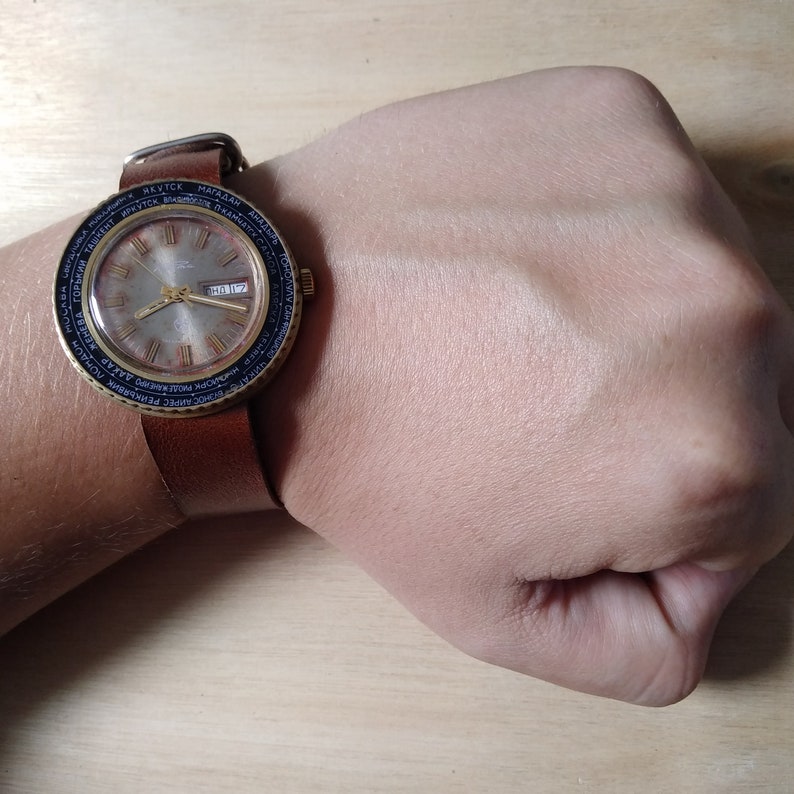 Puede incluir: Un reloj de pulsera de cuero marr&oacute;n con una esfera dorada y marcas negras. La esfera del reloj tiene una ventana de fecha y una pantalla de hora mundial. El reloj est&aacute; en la mu&ntilde;eca de una persona.