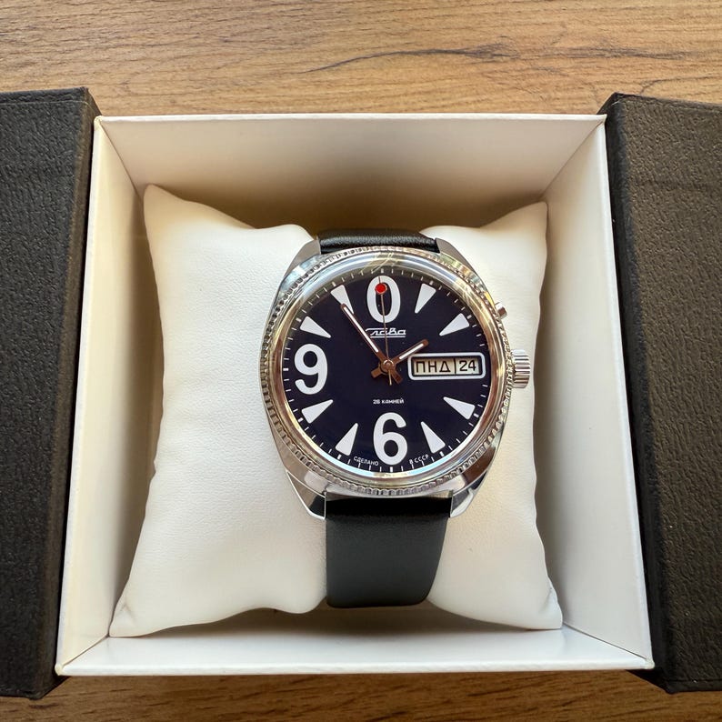 May include: A round wristwatch with a dark blue face, large white numerals, and a black leather band. The watch is in a white box with a black exterior. The watch face has the text "24" and Cyrillic text.