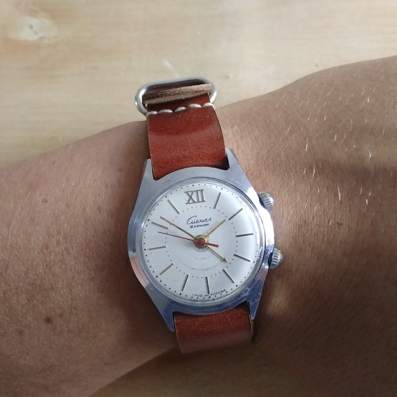 Op de afbeelding: Een zilveren polshorloge met een witte wijzerplaat en zwarte Romeinse cijfers. Het horloge heeft een bruin leren bandje en op de wijzerplaat staat "Слава 18 jewels".