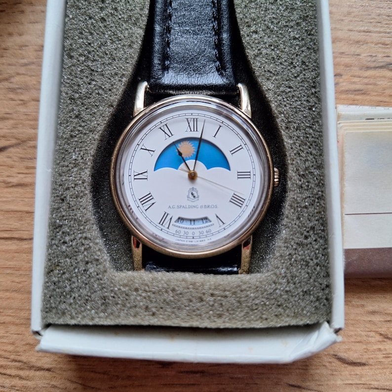 Op de afbeelding: Een goudkleurig horloge met een witte wijzerplaat, Romeinse cijfers en een zwarte leren band. De wijzerplaat heeft een blauw en geel ontwerp en de tekst "A.G. Spalding & Bros."