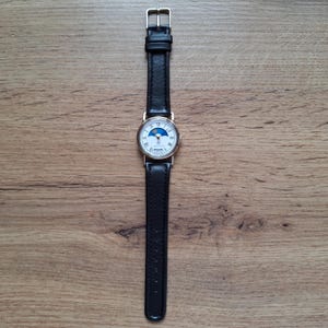 Op de afbeelding: Een goudkleurig polshorloge met een witte wijzerplaat met Romeinse cijfers en een blauw maansikkelontwerp. Het heeft een zwarte leren band en een gouden gesp. Het horloge ligt op een houten oppervlak.