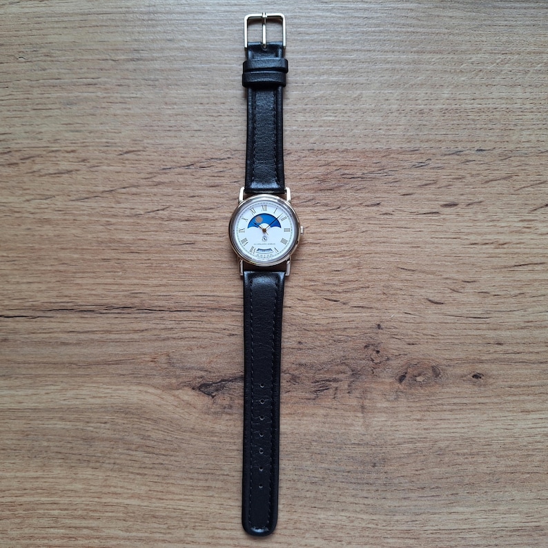 Op de afbeelding: Een goudkleurig polshorloge met een witte wijzerplaat met Romeinse cijfers en een blauw maansikkelontwerp. Het heeft een zwarte leren band en een gouden gesp. Het horloge ligt op een houten oppervlak.