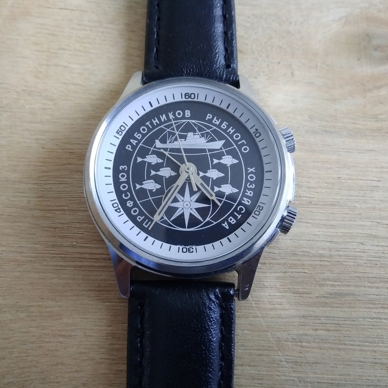 Puede incluir: Un reloj de pulsera plateado con una esfera negra y una correa de cuero negra. La esfera del reloj presenta una br&uacute;jula y un barco con el texto "ПРОФСОЮЗ РАБОТНИКОВ РЫБНОГО ХОЗЯЙСТВА" alrededor del borde.