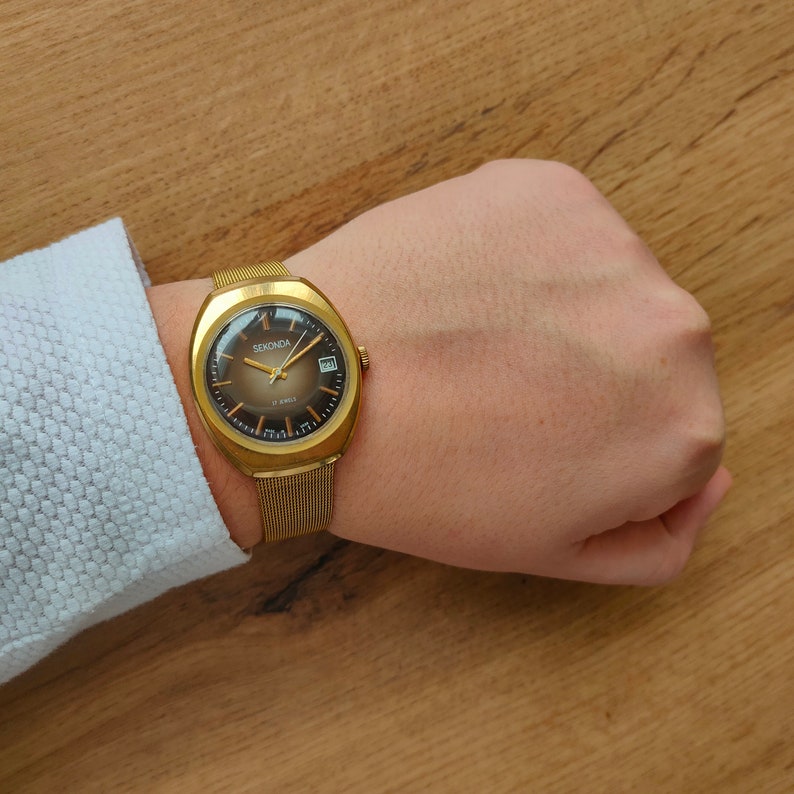 Puede incluir: Un reloj Sekonda dorado con esfera marr&oacute;n y visualizaci&oacute;n de fecha. El reloj tiene una correa de malla y se lleva en la mu&ntilde;eca. La esfera del reloj tiene marcadores y agujas doradas. El reloj est&aacute; sobre un fondo de madera.
