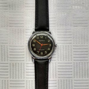 Pu&ograve; includere: Un orologio da polso argentato con un cinturino in pelle nera. Il quadrante dell'orologio &egrave; nero con numeri bianchi e una lancetta dei secondi rossa. Il quadrante dell'orologio ha anche il testo "Кировские" in scrittura cirillica.