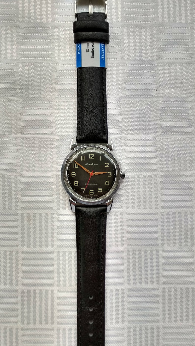 Pu&ograve; includere: Un orologio da polso argentato con un cinturino in pelle nera. Il quadrante dell'orologio &egrave; nero con numeri bianchi e una lancetta dei secondi rossa. Il quadrante dell'orologio ha anche il testo "Кировские" in scrittura cirillica.