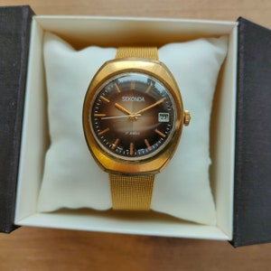 Puede incluir: Un reloj Sekonda dorado con esfera marr&oacute;n y correa de malla. La esfera muestra el nombre de la marca, la fecha y "17 Jewels". El reloj se presenta en una almohada blanca dentro de una caja negra.