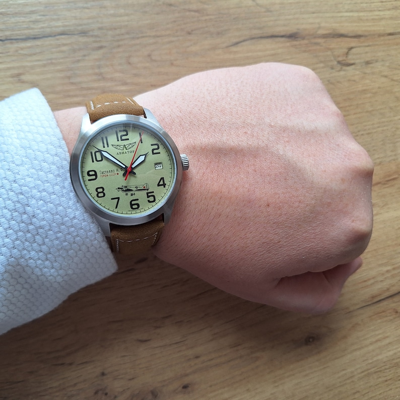 K&ouml;nnte beinhalten: Eine silberne Armbanduhr mit einem braunen Lederarmband. Das Zifferblatt der Uhr ist cremefarben mit schwarzen arabischen Ziffern und einem kleinen Datumsfenster. Die Uhr hat einen roten Sekundenzeiger und eine kleine Flugzeuggrafik auf dem Zifferblatt. Das Zifferblatt der Uhr tr&auml;gt auch den Text "Aviator" und "СССР-1941" in kyrillischer Schrift.