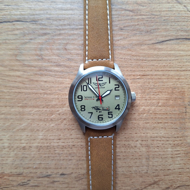 K&ouml;nnte beinhalten: Eine silberne Armbanduhr mit einem braunen Lederband und einem cremefarbenen Zifferblatt. Das Zifferblatt hat schwarze Zahlen und eine kleine Flugzeuggrafik. Das Zifferblatt der Uhr hat auch den Text "Авиатор" und "Гастелло Н.С. 1941 СССР".