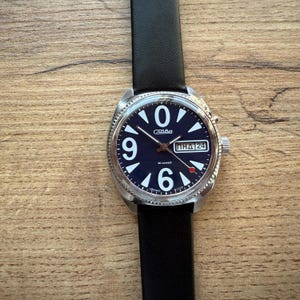 May include: A vintage wristwatch with a dark blue face, featuring white numerals and hands. The watch has a stainless steel case and a black leather strap. The date is displayed in a small window. The watch is on a wooden surface.
