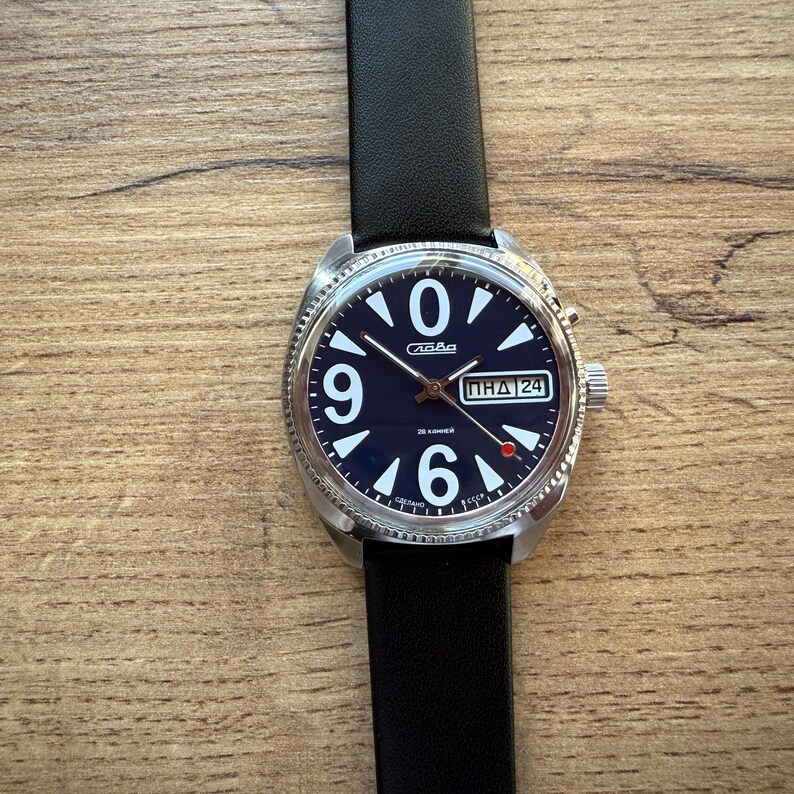 May include: A vintage wristwatch with a dark blue face, featuring white numerals and hands. The watch has a stainless steel case and a black leather strap. The date is displayed in a small window. The watch is on a wooden surface.