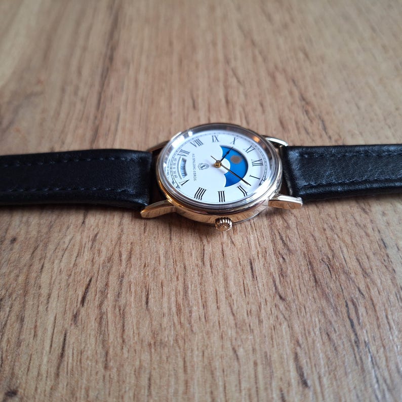 Op de afbeelding: Een goudkleurig polshorloge met een witte wijzerplaat, Romeinse cijfers en een blauwe maanfase-indicator. Het horloge heeft een zwarte leren band. Het horloge ligt op een houten oppervlak.