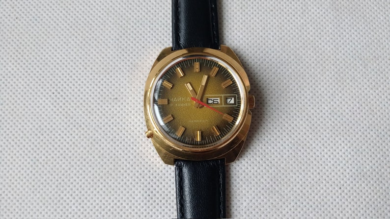 May include: A gold-toned wristwatch with a black leather band. The watch face is green with gold-colored hour markers and hands. The watch has a date window at the 3 o'clock position and the text "Чайка 24 камня" is visible on the watch face.