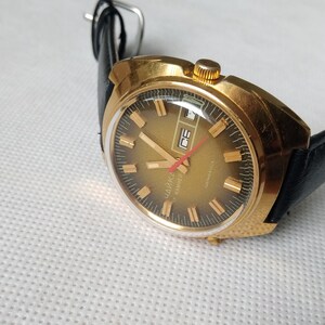 May include: A gold-toned wristwatch with a black leather band. The watch face has a black background with gold-colored hour markers and hands. The watch has a date window at the 3 o'clock position. The watch is made by the brand "SLAVA" and says "Automatic" on the face.