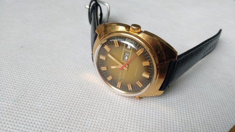 May include: A gold-toned wristwatch with a black leather band. The watch face has a black background with gold-colored hour markers and hands. The watch has a date window at the 3 o'clock position. The watch is made by the brand "SLAVA" and says "Automatic" on the face.