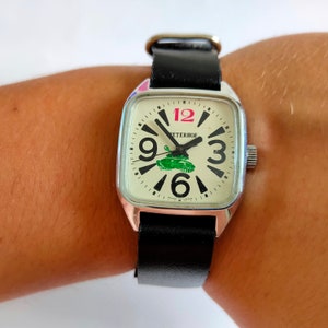 Puede incluir: Un reloj de pulsera cuadrado con una correa de cuero negra. La esfera del reloj es blanca con números y manecillas negros. Un tanque verde está impreso en la esfera del reloj con la palabra "PETERHOF" encima. El reloj tiene una caja plateada y un 12 rosa.