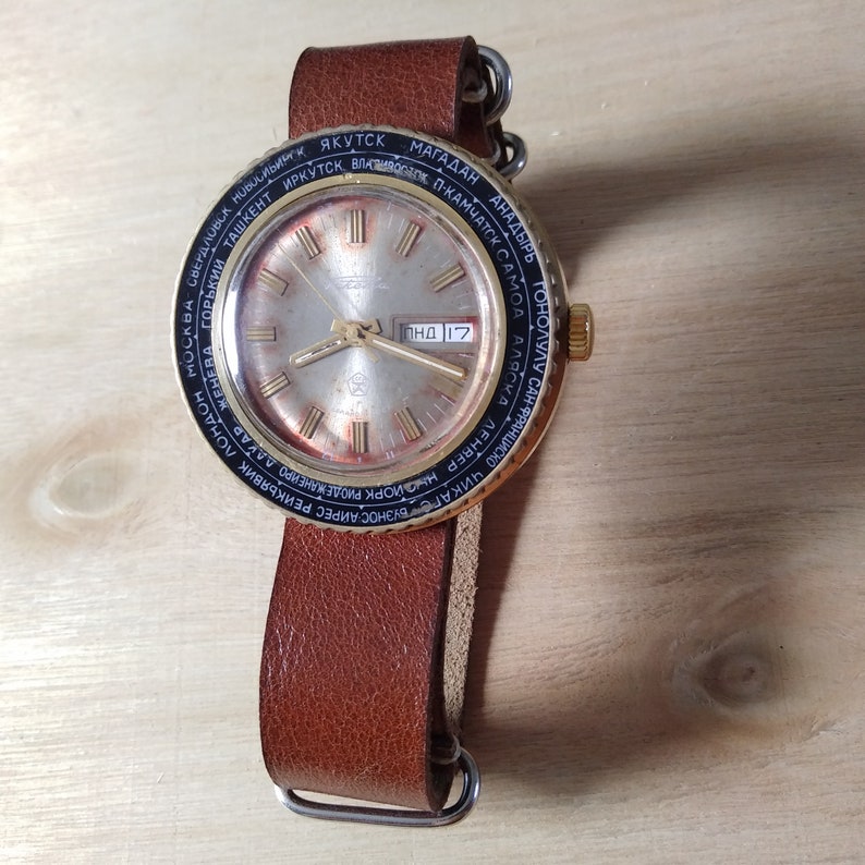 Puede incluir: Un reloj de pulsera dorado con una esfera negra y una correa de cuero marr&oacute;n. La esfera del reloj tiene una esfera blanca con marcadores de hora romanos negros y una ventana de fecha. El reloj tiene un bisel de hora mundial con nombres de ciudades impresos en negro.