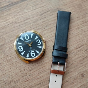 May include: A gold-tone wristwatch with a black face and white numerals. The watch face says "PAKEТА" and "СДЕЛАНО В СССР". The watch is on a black leather strap.