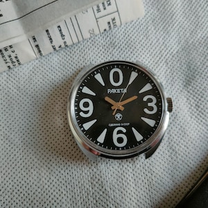 May include: A vintage Raketa watch face with a black dial and white numerals. The watch face features the word "PAKETA" and the text "MADE IN USSR". The hands are gold-coloured, and the watch has a silver-coloured bezel.
