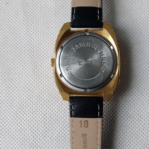 May include: The back of a gold-tone wristwatch with a black leather band. The watch face is covered with a metal plate that says "ЭЛЕКТРОННЫЕ ЧАСЫ" in Cyrillic lettering.