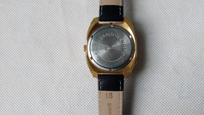 May include: The back of a gold-tone wristwatch with a black leather band. The watch face is covered with a metal plate that says "ЭЛЕКТРОННЫЕ ЧАСЫ" in Cyrillic lettering.