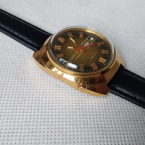May include: A gold-tone wristwatch with a black leather band. The watch face is green with gold-tone Roman numeral hour markers and a date window at the 3 o'clock position.
