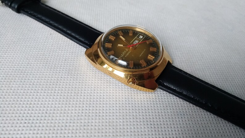 May include: A gold-tone wristwatch with a black leather band. The watch face is green with gold-tone Roman numeral hour markers and a date window at the 3 o'clock position.