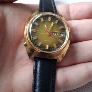 May include: A gold-tone wristwatch with a black leather band. The watch face is green with gold-tone markings and a date window. The watch says "Чайка" in Cyrillic letters.