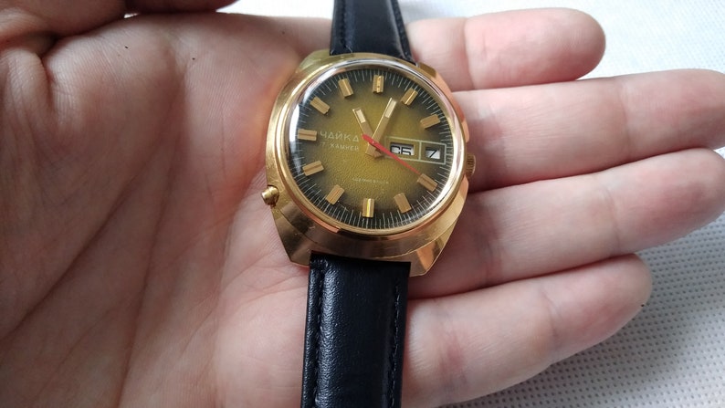 May include: A gold-tone wristwatch with a black leather band. The watch face is green with gold-tone markings and a date window. The watch says "Чайка" in Cyrillic letters.