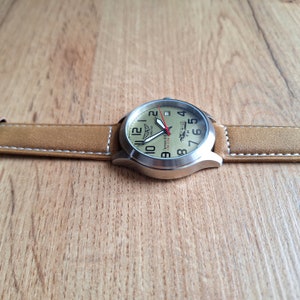 K&ouml;nnte beinhalten: Eine silberne Armbanduhr mit einem braunen Lederarmband. Das Zifferblatt der Uhr ist cremefarben mit schwarzen Zahlen und einem roten Sekundenzeiger. Das Zifferblatt der Uhr zeigt auch eine kleine Flugzeuggrafik und den Text "Action Pilot".