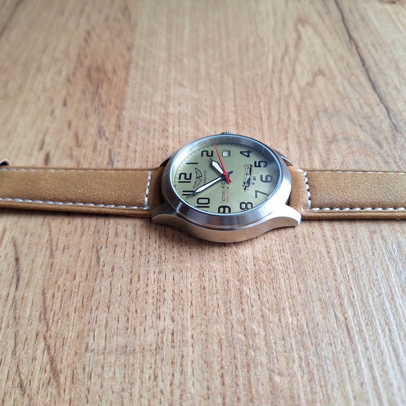 K&ouml;nnte beinhalten: Eine silberne Armbanduhr mit einem braunen Lederarmband. Das Zifferblatt der Uhr ist cremefarben mit schwarzen Zahlen und einem roten Sekundenzeiger. Das Zifferblatt der Uhr zeigt auch eine kleine Flugzeuggrafik und den Text "Action Pilot".