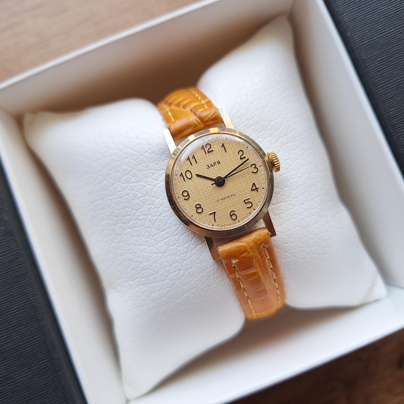 Op de afbeelding: Een vintage goudkleurig polshorloge met een lichtbruine leren band, gepresenteerd in een witte doos. De wijzerplaat toont zwarte cijfers en wijzers tegen een lichtgouden achtergrond. De band is ongeveer 1,5 cm breed.