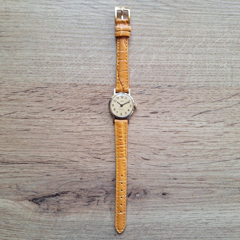 Op de afbeelding: Een vintage polshorloge met een lichtbruine leren band en een goudkleurige gesp. De wijzerplaat is cr&egrave;mekleurig met zwarte cijfers en wijzers. Het horloge ligt op een houten achtergrond.