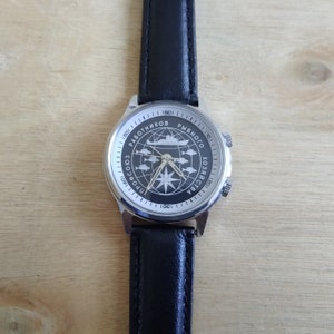 Puede incluir: Un reloj de pulsera plateado con una correa de cuero negro. La esfera del reloj tiene un fondo negro con una br&uacute;jula blanca y un barco con el texto "ПРОФСОЮЗ РАБОТНИКОВ РЫБНОГО ХОЗЯЙСТВА 1901-1984" alrededor de la br&uacute;jula.