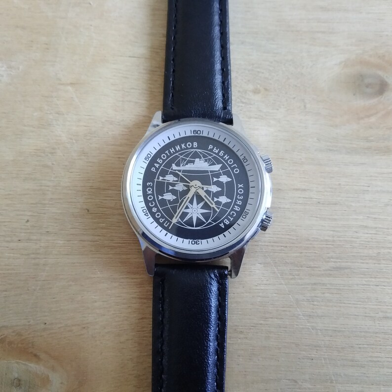 Puede incluir: Un reloj de pulsera plateado con una correa de cuero negro. La esfera del reloj tiene un fondo negro con una br&uacute;jula blanca y un barco con el texto "ПРОФСОЮЗ РАБОТНИКОВ РЫБНОГО ХОЗЯЙСТВА 1901-1984" alrededor de la br&uacute;jula.