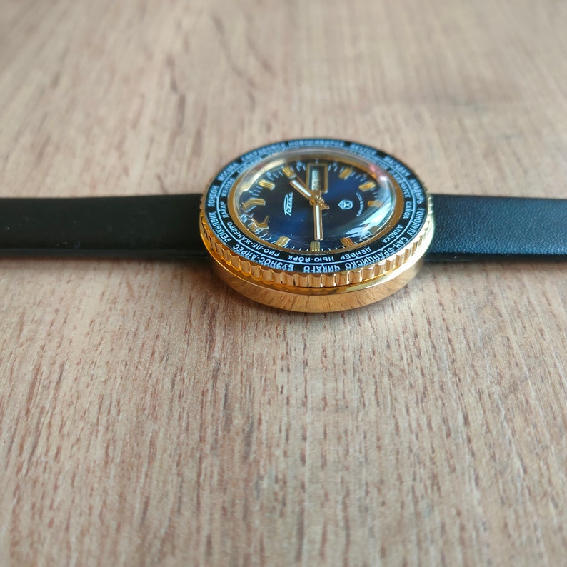 May include: A gold-toned wristwatch with a black face and a black leather band. The watch face has a world time bezel with city names printed in black and white.
