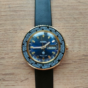 May include: A gold-toned wristwatch with a blue face and a black leather band. The watch face features a world time display with city names around the edge. The date window displays "СРД 118".