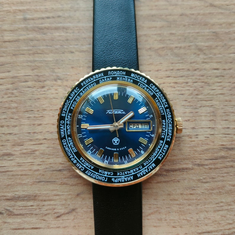 May include: A gold-toned wristwatch with a blue face and a black leather band. The watch face features a world time display with city names around the edge. The date window displays "СРД 118".