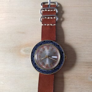 Puede incluir: Un reloj de pulsera dorado con una esfera negra y una correa de cuero marr&oacute;n. La esfera del reloj tiene un dial de hora mundial con nombres de ciudades impresos alrededor del borde. La ventana de fecha muestra "ЧНД 17".