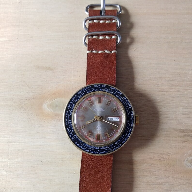 Puede incluir: Un reloj de pulsera dorado con una esfera negra y una correa de cuero marr&oacute;n. La esfera del reloj tiene un dial de hora mundial con nombres de ciudades impresos alrededor del borde. La ventana de fecha muestra "ЧНД 17".